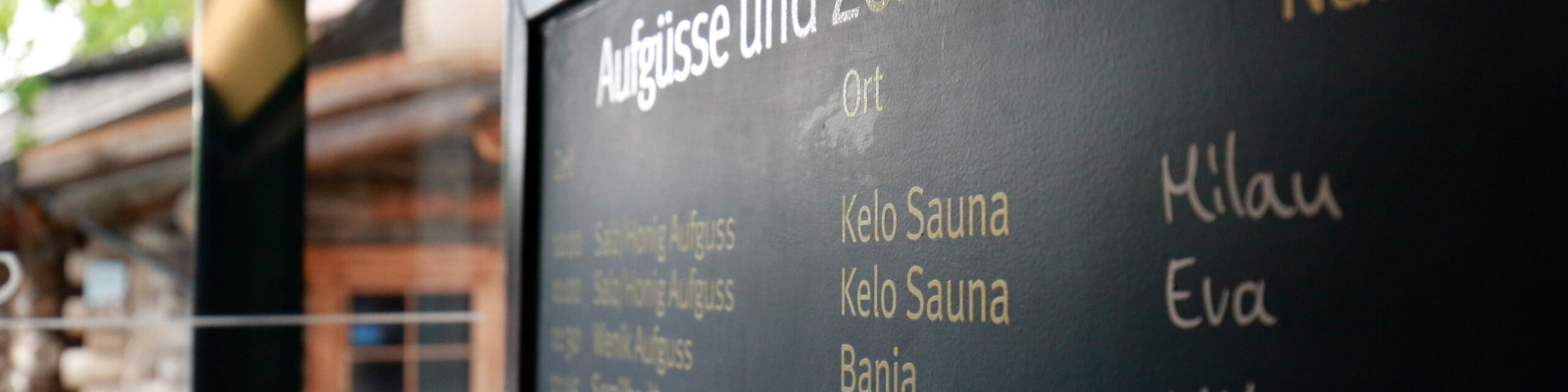 Ein Ausschnitt einer Tafel auf der Aufgüsse, Sauna und Mitarbeitender stehen. Im Hintergrund ist das Saunadorf sichtbar