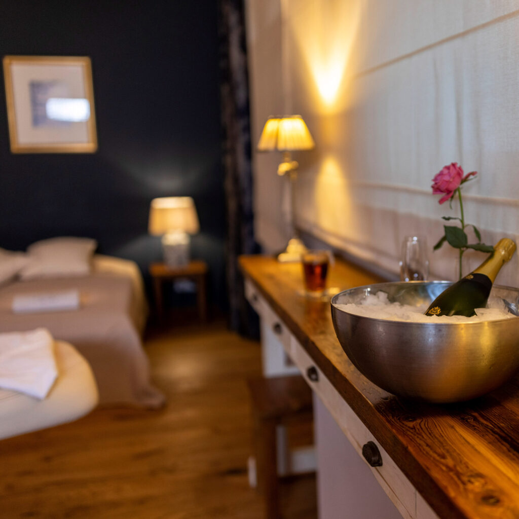 Ein Sideboard aus Holz mit einem Champagnerkühler und einer roten Rose. Im Hintergrund ist ein Bett zu sehen.