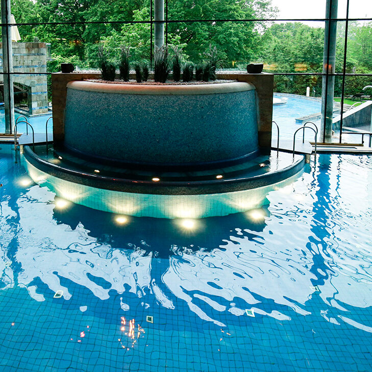 Blick in ein großes Schwimmbecken. Im Hintergrund wird durch eine Glasfront ein Außenpool in einem grünen Garten sichtbar. Die Sonne scheint.