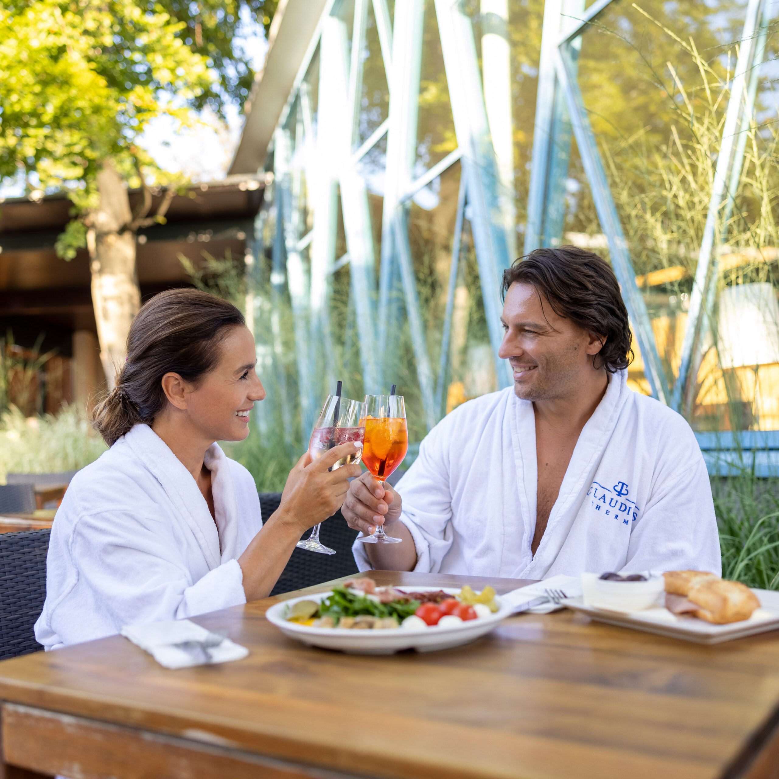 Ein lachender Mann und eine lachende Frau sitzen in weißen Bademänteln im Außenbereich an einem Holztisch. Sie Stoßen mit Getränken an. Vor ihnen stehen zwei Teller mit Essen.