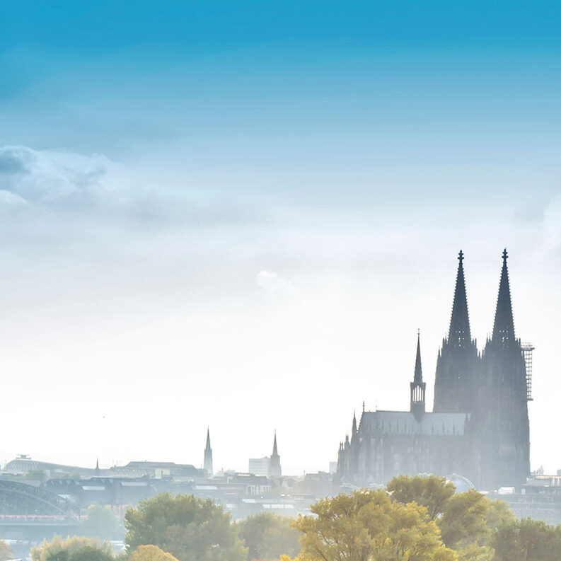 Im Vordergrund ist grüne Wiese und Büsche und Bäume in Herbstfarben. Im Hintergrund sieht man die Skyline von Köln, in der Mitte den Kölner Dom.