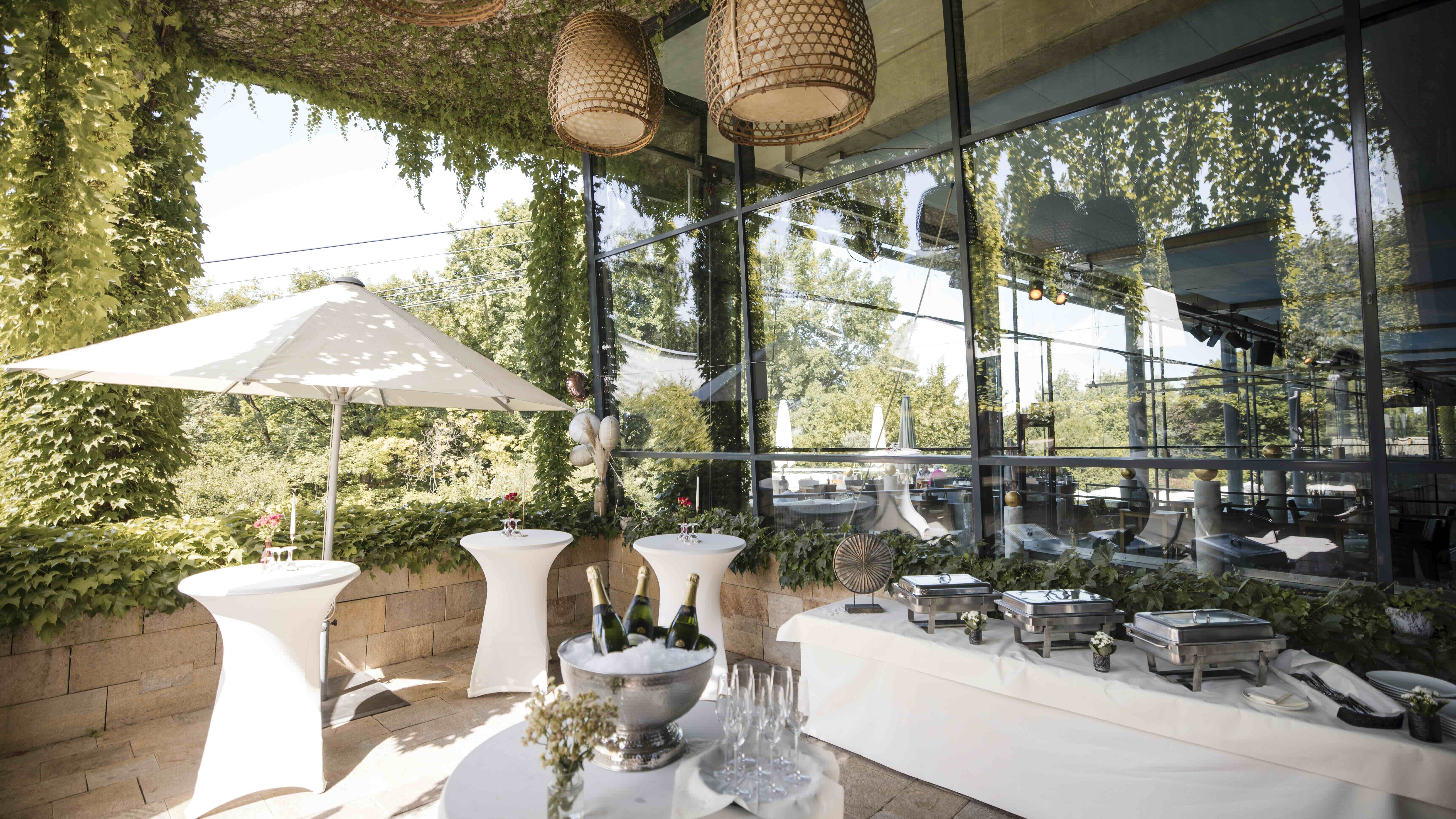 Terrasse mit viel Grün und einem Sonnenschirm. Ein Buffet ist aufgebaut und einige Stehtische