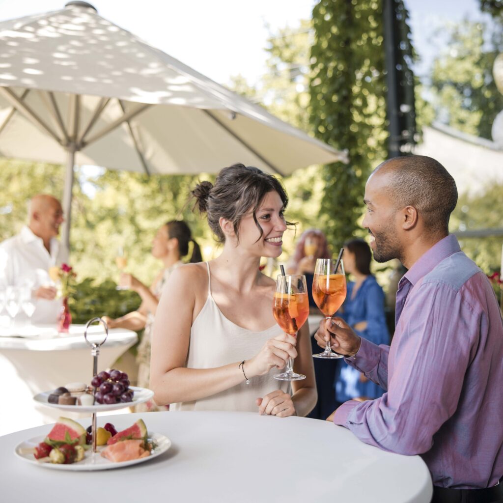 Ein junges Paar stößt mit einem orangenen Aperitiv an. Es steht auf einer sonnigen Terrasse mit einem weißen Sonnenschirm und viel Grün im Hintergrund