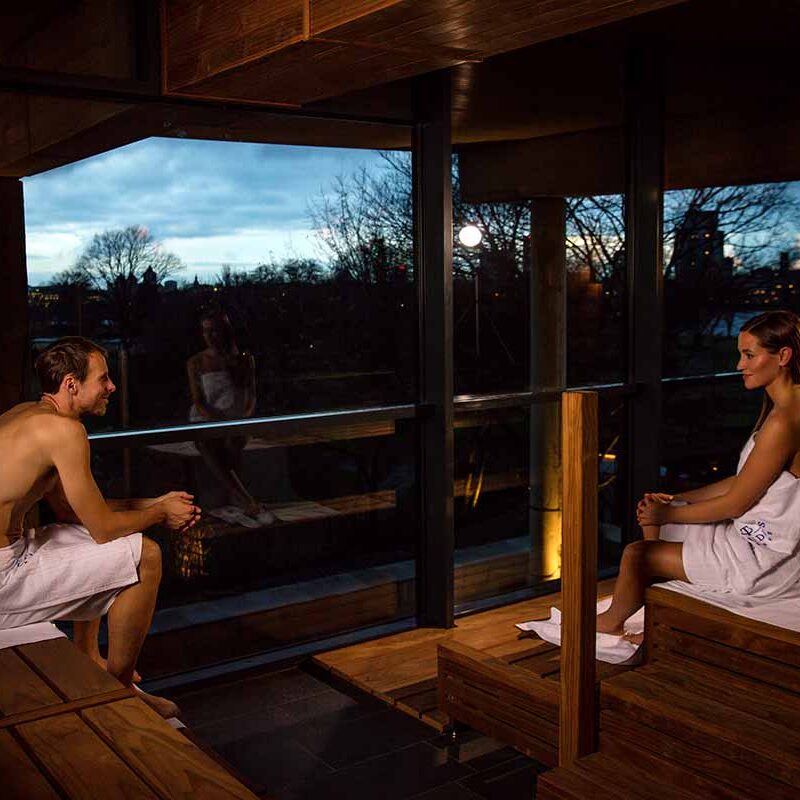 Zwei Personen sitzen in Handtüchern auf Holzbänken einer Sauna mit großer Glasfront. Draußen ist Abenddämmerung, Bäume und die Silhouette einer Kirche sind im Hintergrund zu sehen. Die beiden sitzen sich entspannt gegenüber und sprechen miteinander.