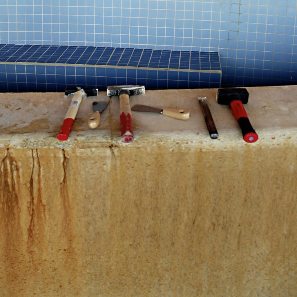 Verschiedene Hammer und Spachtel liegen auf einer ungefließten Mauer. Dahinter ist eine Mauer mit quadratischen, blauen Fliesen zu sehen.