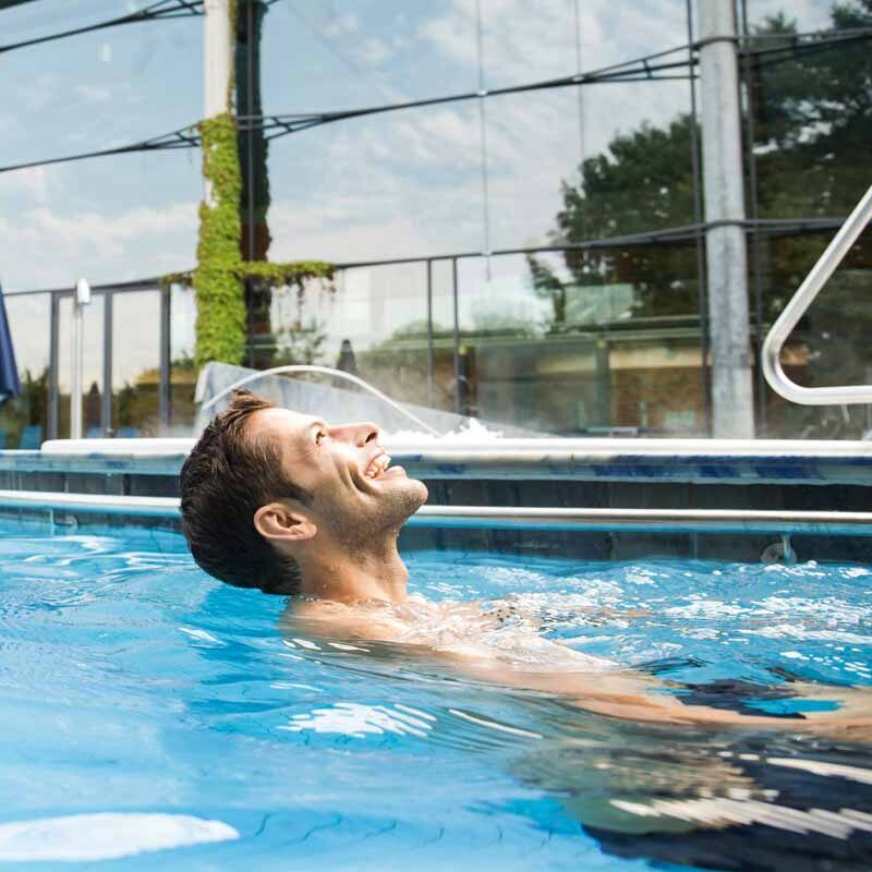 Ein Mann entspannt im Außenbecken einer Therme und lehnt sich lächelnd im warmen Wasser zurück. Hinter ihm spiegeln sich Bäume und Himmel in den großen Glasfassaden des Gebäudes, daneben stehen geschlossene Sonnenschirme